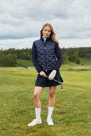 Girl wearing a navy jacket and navy skort is standing on a golf course and holding a golf club.