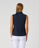 A woman viewed from the back wearing a sleeveless navy equestrian polo shirt and white riding pants, standing against a light gray studio background.