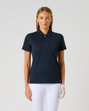 A woman modeling a short-sleeve polo shirt with a half-zip collar in dark navy blue, paired with white pants, against a light gray studio background.