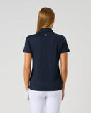 A woman viewed from the back wearing a dark navy short-sleeve polo shirt and white pants, standing against a plain light gray studio background.
