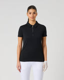 A woman modeling a black short-sleeve polo shirt with a half-zip collar, paired with white pants, against a light gray studio background.