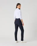 A woman stands in profile wearing a white long-sleeve mock-neck top, dark navy fitted trousers, and white sneakers against a plain light gray studio background.