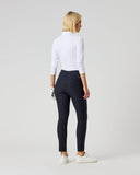A woman with blonde hair is shown from behind wearing a white long-sleeve mock neck top, dark navy high-waisted riding tights, and white sneakers, standing against a light gray studio background.