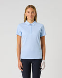 A woman modeling a light blue short-sleeve polo shirt with a half-placket collar, paired with dark pants and a white golf glove, against a plain light gray studio background.