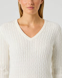 A woman wearing a cream-colored cable-knit V-neck sweater against a light gray studio background.