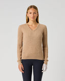 A woman modeling a beige ribbed V-neck sweater with long sleeves, paired with dark pants and white gloves, against a plain light gray studio background.
