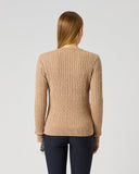 A woman viewed from behind wearing a beige cable-knit sweater and dark navy pants, standing against a plain light gray studio background.