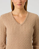 A woman wearing a beige cable-knit V-neck sweater, shown from the nose down against a light gray studio background.