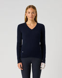 A woman stands against a light gray studio background wearing a dark navy ribbed V-neck sweater, dark navy leggings, and white gloves.