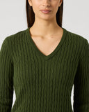A woman wearing a dark olive green cable-knit V-neck sweater, shown from the chest up against a light gray studio background.