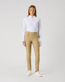 A woman stands against a light gray studio background wearing a white long-sleeve collared shirt, tan slim-fit trousers, and white sneakers.