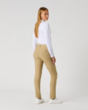 A woman stands in profile wearing slim-fit beige trousers, a white long-sleeve top, and white sneakers against a plain light gray studio background.