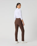 A woman wearing brown slim-fit trousers, a white long-sleeve top, and white sneakers, standing in profile against a light gray studio background.