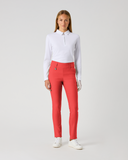 A woman modeling bright coral-red slim-fit pants with a white long-sleeve polo shirt and white sneakers, standing against a light gray studio background.