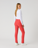A woman standing in profile wearing bright coral-red slim-fit pants, a white long-sleeve top, and white sneakers against a light gray studio background.