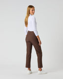 A woman stands in profile wearing brown straight-leg trousers, a white long-sleeve top, and white sneakers against a light gray studio background.
