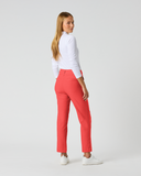 A woman modeling bright coral-red straight-leg trousers with a white long-sleeve top and white sneakers, shown from a side-back angle against a light gray studio background.