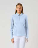 A woman modeling Daily Sports light blue long-sleeve Peoria polo shirt with a half-zip collar, paired with white pants, against a solid light gray background.