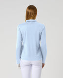 A woman viewed from behind wearing a light blue long-sleeve knit top and white pants against a plain light gray studio background.