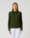 A woman modeling a dark olive green long-sleeve polo shirt with a half-zip collar, paired with white pants, against a solid light gray background.