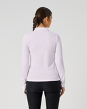 A woman viewed from behind wearing a light lavender long-sleeve athletic top and black leggings against a plain light gray studio background.