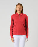 A woman modeling a red long-sleeve polo shirt with a half-zip collar, paired with white pants, against a solid light gray background.