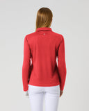 A woman viewed from behind wearing a bright red long-sleeve polo shirt and white pants against a light gray studio background.