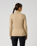 A woman viewed from the back wearing a beige ribbed mock-neck long-sleeve top and black pants, standing against a plain light gray studio background.