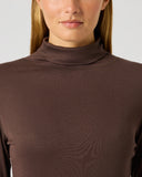 A close-up of a woman wearing a ribbed, long-sleeve mock neck turtleneck in a solid dark brown color, set against a plain light gray background.