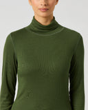A woman wearing a dark olive green ribbed long-sleeve turtleneck top, shown from the nose down against a light gray studio background.