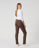 A woman stands in profile wearing slim-fit brown pants, a white long-sleeve top, and white sneakers against a plain light gray studio background.
