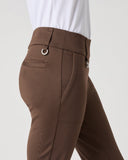 A close-up side view of a person wearing brown tailored riding pants with a white top, shown against a plain light gray studio background.
