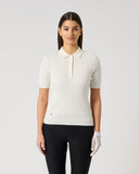 A woman modeling a short-sleeve ribbed knit polo shirt in off-white, paired with black pants and a white glove, against a plain light gray studio background.