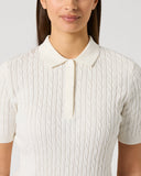 A woman wearing a short-sleeve polo shirt with a cable-knit texture in off-white/cream, shown from the nose down against a light gray studio background.
