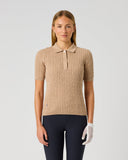 A woman modeling a beige ribbed short-sleeve polo shirt with a three-button placket, paired with dark navy pants and a white golf glove, against a light gray studio background.