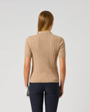 A woman viewed from behind wearing a beige short-sleeve ribbed knit polo shirt and navy blue pants, standing against a plain light gray studio background.