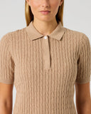 A woman wearing a beige short-sleeve knit polo shirt with a cable pattern, shown from the chest up against a light gray studio background.