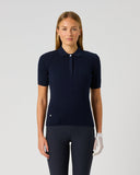 A woman stands against a light gray studio background wearing a dark navy ribbed short-sleeve polo shirt with a small white button and matching dark navy pants. She is also wearing a white golf glove.