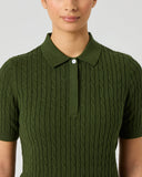 A woman wearing a dark olive green short-sleeve polo shirt with a cable-knit texture, shown from the chest up against a light gray studio background.