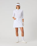 A woman modeling a white golf outfit consisting of a long-sleeve collared shirt, a short skirt, a visor, gloves, socks, and shoes, standing against a light gray studio background.