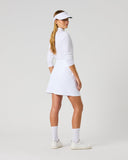 A woman modeling a white long-sleeve tennis dress, white visor, white socks, and white sneakers, shown from a side-back angle against a light gray studio background.
