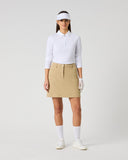 A model stands against a light gray studio background wearing a white long-sleeve polo shirt, a beige mini skirt, white socks, white shoes, a white visor, and white gloves.
