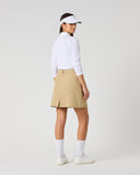 A woman stands in profile wearing a white long-sleeve athletic top, a beige golf skirt, white socks, and white sneakers against a light gray studio background.