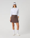 A model stands against a light gray studio background wearing a white long-sleeve collared polo shirt, a brown mini skirt, white socks, white shoes, a white visor, and white gloves.