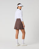 A woman stands in profile wearing a white long-sleeve athletic top, a brown pleated tennis skirt, white socks, and white sneakers against a light gray studio background.