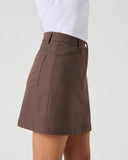 A person wearing a brown A-line mini skirt with a side pocket, paired with a white top, shown from the waist down against a light gray studio background.