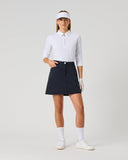A woman modeling a white long-sleeve polo shirt, a dark navy blue golf skirt with white side stripes, a white visor, white gloves, white socks, and white sneakers, standing against a light gray studio background.