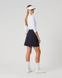 A woman modeling a white long-sleeve golf shirt, navy blue pleated golf skirt, white visor, white socks, and white golf shoes, shown from a side-back angle against a light gray studio background.