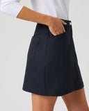 A woman wearing a dark navy blue A-line mini skirt with side pockets, paired with a white long-sleeve top, shown from the waist down against a light gray studio background.
