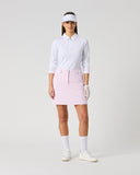 A model stands against a light gray studio background wearing a white long-sleeve collared golf shirt, a light pink mini skirt, white socks, white shoes, a white visor, and white gloves.